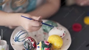 A close up video of the egg painting process. A little girl paints a ceramic egg. She uses a brush in order to make unequal dots or red colour. The background is blurred.