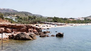 Sun-kissed Greek Coastal Scene, Rocky Outcrops Meet Crystal-clear Blue Waters, While a Sandy Beach Teems With People and Colorful Umbrellas. Vacation Vibes