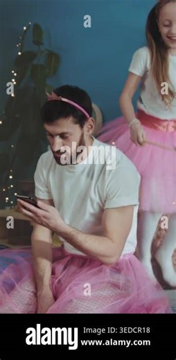 caucasian father scrolling phone while daughter twirls in pink tutu in living room string lights and makeshift stage Stock Video Footage - Alamy