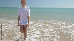 Pretty smiling teenage girl in a T-shirt walks at ocean shore, sea surf at sunrise. Young caucasian girl going along empty seaside, in the morning. Happy lady relaxing at vacation