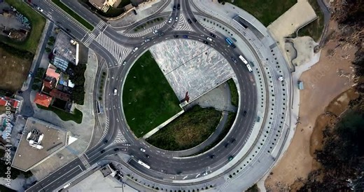 Top-down aerial view of a coastal roundabout in Porto, Portugal, showing flowing traffic, road markings, nearby buildings, green space, and the Atlantic shoreline.