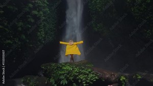 Blonde girl at waterfall flowing in Bali raises hands up. Hiking, Woman in a yellow raincoat or jacket in the tropical jungle enjoys a vacation or trip, lifts palms. Rainy season, Indonesia.