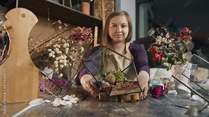 Woman shows off a beautifully crafted floral arrangement. The arrangement was created by a skilled craftswoman using succulents, decorative stones, and sand, and it is presented in a sleek glass