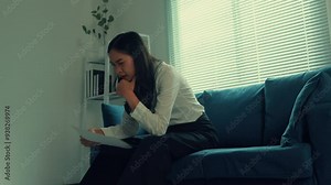 Businesswoman sitting on a blue sofa reads a document, breaking into tears upon realizing the bad news. Emotions of sadness and despair overwhelm her as she processes the situation