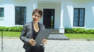 Female real estate agent smiling at the camera while holding a clipboard and standing in from of a modern house to be sell. Shot in 4k resolution