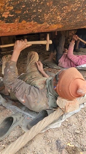 Lying under the ship, he is using a hammer to remove rust and paint from a large ship.