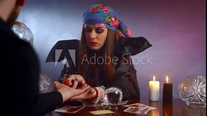 Young fortune teller reading fortune lines on male palm or hand, mystic esoteric or paranormal chiromancy concept. Future prediction with crystal ball and burning candles in dark room. Symbolism in
