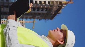 Vertical Screen: Building inspector wearing hard hat and safety vest making notes on paper and talking, multistory houses on background. Low angle man inspecting construction site. Concept of control