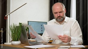 Confused businessman having difficulty with paperwork while sitting at workplace. Entrepreneur looks at documents, spreads his hands and cannot understand problem. Male professional working on laptop