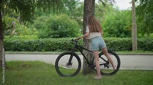 Woman in jean shorts approaches bicycle leaning against tree in serene park, removes stand, and begins moving with bike, background of lush greenery and paved path