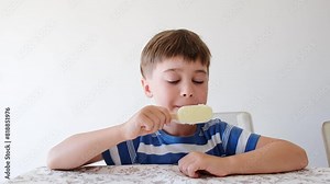 Handsome boy eating popsicle on a stick. He bites off the ice cream with pleasure. Close-up. Child eat in the kitchen or at the dining room table. Tasty dessert. Slow motion. High quality 4k footage