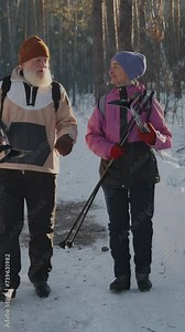 Full vertical tracking shot elderly Caucasian couple, wearing warm snow suits, walking through snowy forest, carrying cross-country skis, chatting, man telling story and woman reacting with surprise