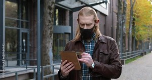 Handsome ginger man in protective mask paying online with a credit card number on the tablet while shopping online and standing at the street. Outdoors