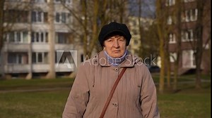 Elderly woman stands outside and looks straight. Soviet style of grandmother clothes. Early spring walk. Sad, lonely granny. Wrinkled woman face. Old apartment buildings in the background.