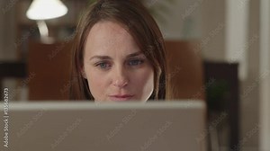 Young woman watches videos on internet in her laptop at home. She watches very concentrated and serious. Dolly shot parallel to the woman. Extreme close up with the laptop at foreground.