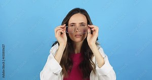 Girl with poor vision looking at camera and rejoicing after putting on eyeglasses isolated against blue background. Young brunette woman with vision problems seeing perfectly with eyeglasses. Try see.