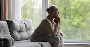 Side view 65s widow lonely grey-haired woman sits on sofa in living room looking into distance, feeling solitude, recollects past, missing longing for husband. Retiree in nursing home, ageing concept