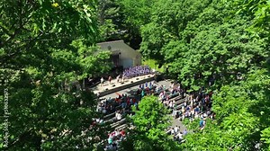 Aerial framing among trees of outdoor concert. Crowd of people gather in park for music.