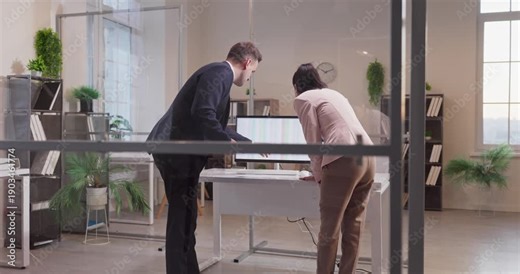 Business colleagues in office meeting around computer. A businessman and businesswoman lean over a desk reviewing data on monitors, engaged in discussion. Clear teamwork and collaboration theme.