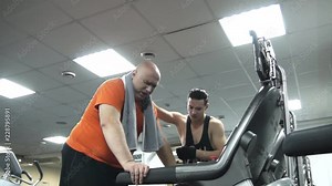 Fat man is very tired on the treadmill breathing hardly. His young coach motivates him, forsec to go on, explains. The view from the bottom up