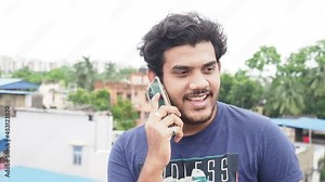 Handsome Indian guy calling someone from his smartphone while sitting in a terrace. Young attractive man in casual wear,happily talking to his friend over a phone call - lifestyle, technology concept.