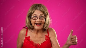 Funny elderly mature woman, 80s, wearing negligee and glasses giving thumbs up hand gesture, smiling and walking across pink background.