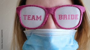 Woman put on a face mask to celebrate at a bachelorette party. Young bride wearing pink glitter sunglasses pulls up a blue medical mask. Represents wedding showers and parties cancelled from Covid-19