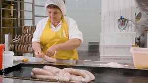 Woman worker in uniform butcher processing raw sausages fastening ends with ropes on meat factory. Production line of meat products on food plant. Manual labor, hand work. Food sausage manufacturing.