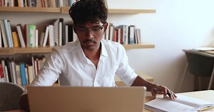 Serious concentrated Indian student guy e-learning theory, reading subject topics, gains new knowledge, improve skills, making notes, writing exercise, doing test using laptop app, studying in library
