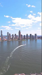 erial video of the Hudson River on a sunny day in New York City, showcasing the vibrant skyline and calm waters with boats gliding through, under a bright blue sky dotted with fluffy clouds.