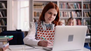 Attractive young red haired girl with freckles preparing for exam and studying at university library. Charming young student focused on learning and make notes in her notebook.
