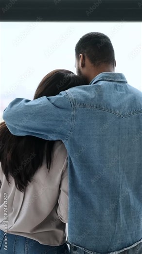 A loving couple embraces while looking out the window of their home, sharing a quiet, intimate moment together. Rear view. Vertical video.