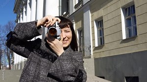 Young woman tourist in vintage gray coat photographing architecture in empire style on retro camera.