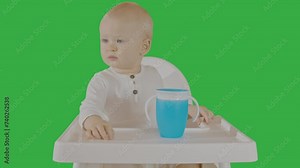 Serious baby boy in high chair with blue cup, early meal time, green screen setup. Cute Infant at feed time, explore sippy cup, child development, chroma key background for visual content creation.