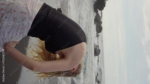 Side view of a blonde woman with long hair that flutters in the wind. A girl walks along the black sand beach of the Ocean. Sensual Girl dressed in a short summer skirt. Vertical video. Slow motion