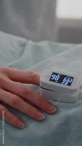 Vertical video: Close up of hospitalized woman with finger heart rate monitor showing pulse while resting in bed during cardiology consultation in hospital ward. Patient with oximeter waiting for