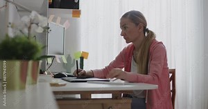 A blond woman is focused on remote work in her home office, surrounded by plants and sticky notes on her monitor.