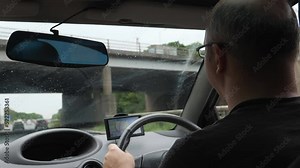 Rear left side view of driver with eyeglasses driving private car on highway in England, using sat nav or GPS, selective focus on man's head.