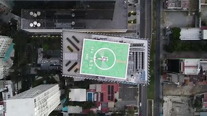 Top down aerial view of a helicopter with rotor spinning on a symmetrical helipad at the top of a skyscraper surrounded by tall buildings of the city. Cinematic shot from a smoothly climbing drone.