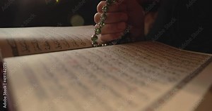 A Muslim person using a prayer beads, with an opened Quran, the focus moves from the prayer beads to the quranic verses