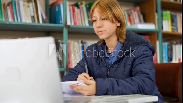 Young student using a computer and taking notes for a school assignment in a public library