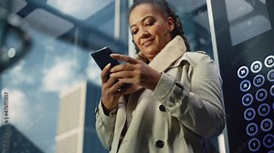 Successful Black Adult Female Riding Glass Elevator to Office in Modern Business Center. Beautiful African American Manager Using Smartphone, Write Message, Check Social Media and Emails in a Lift.