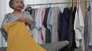 Footage medium shot of Asian Senior Woman in Laundry Room. Authentic real life older lady doing everyday routine, ironing clothes with steam iron and Put t-shirt on a hanger
