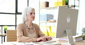 Focused mature woman types on keyboard looking at screen of computer. Serious grey-haired manager at workplace in office slow motion