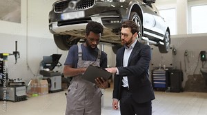 Mechanic explaining car repair details to customer in modern auto shop. Professional interaction between mechanic and client in vehicle service center with car on lift.