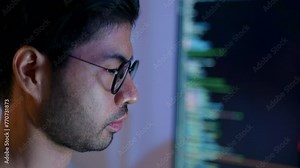 Close-up of a focused male programmer reviewing complex code on a computer monitor in a dimly lit room.