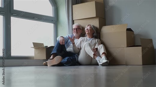 A happy retired couple takes a break from moving into a new flat, relaxing on the floor among cardboard boxes and potted plants while talking and planning interior design together