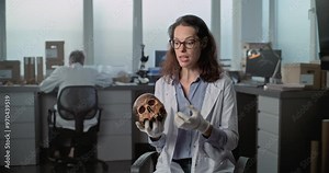 Female scientist speaks to camera, shows fossil skull of prehistoric human during interview for popular science film in archaeological scientific laboratory. Colleague works in background. Static Shot