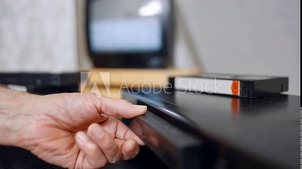 VHS player, several tapes and shimmering screen. A cassette appears from the player and hand puts it on the pile.