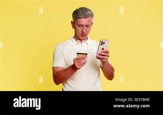 Middle-aged man holding smartphone and credit card while attentively entering payment information to make online purchase shopping. Young guy concentrated on transaction isolated on yellow background Stock Video Footage - Alamy